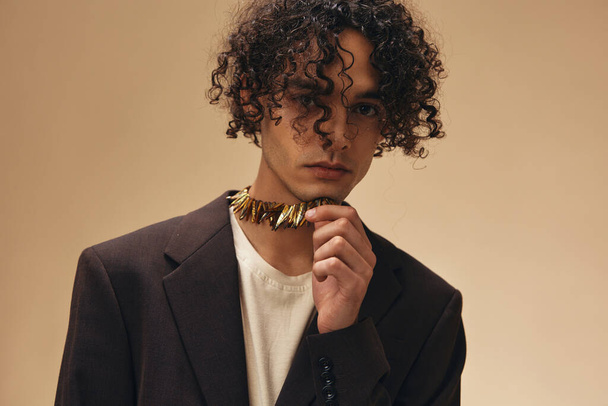 Awesome tanned curly man in classic brown jacket touches ethnic necklace posing isolated on over beige pastel background. Fashion New Collection offer. Retro style concept. Free place for ad - Photo, Image
