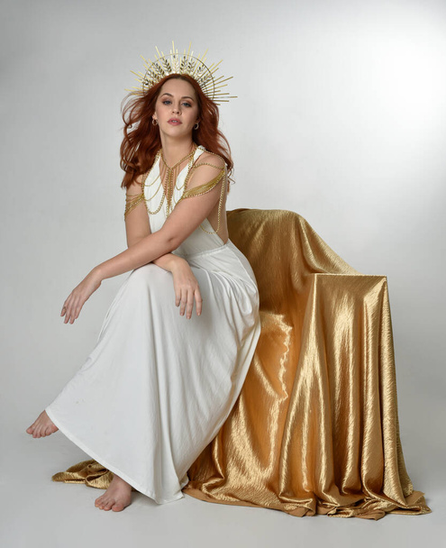 portrait of beautiful red head woman wearing long flowing fantasy toga gown with angelic  golden halo crown on studio background - 写真・画像