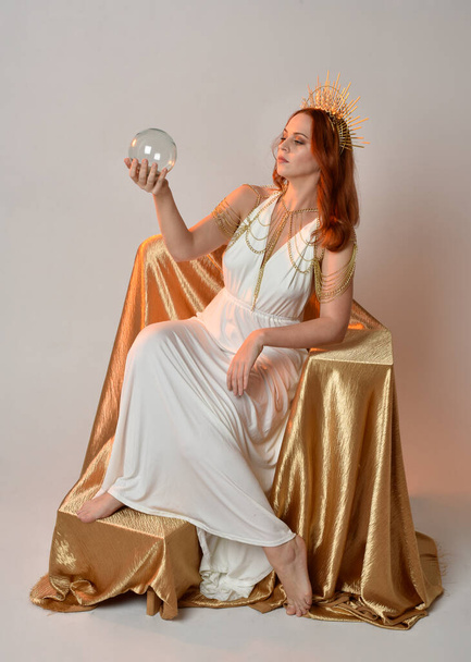 portrait of beautiful red head woman wearing long flowing fantasy toga gown with angelic  golden halo crown on studio background - Valokuva, kuva