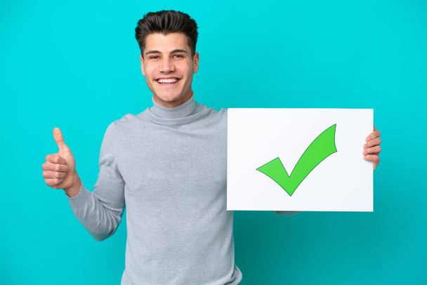 Young handsome caucasian man isolated on blue bakcground holding a placard with text Green check mark icon with thumb up - Photo, Image