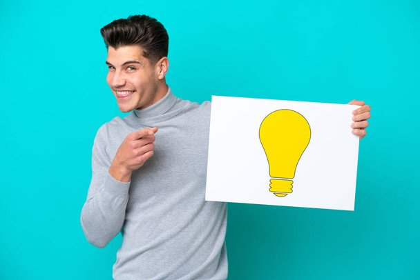 Young handsome caucasian man isolated on blue bakcground holding a placard with bulb icon and pointing to the front - Foto, Imagem