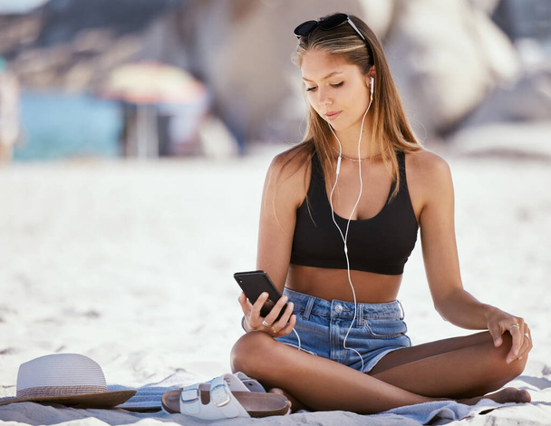 One beautiful young caucasian woman relaxing and listening to music while sitting on the beach. Enjoying a summer vacation or holiday outdoors in the day. Taking time off and getting away from it all. - 写真・画像