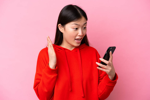 Młoda chińska kobieta odizolowana na różowym tle patrząc w aparat podczas korzystania z telefonu komórkowego z zaskakującym wyrazem - Zdjęcie, obraz