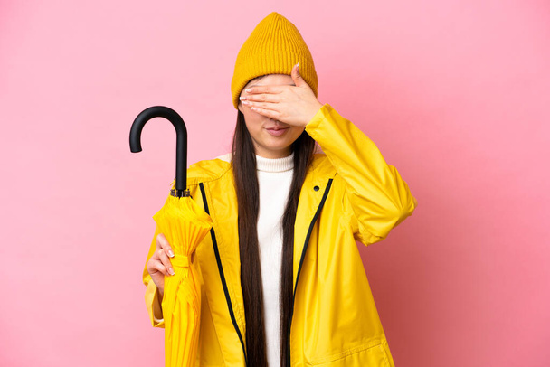 Young Chinese woman with rainproof coat and umbrella isolated on pink background covering eyes by hands. Do not want to see something - Foto, afbeelding