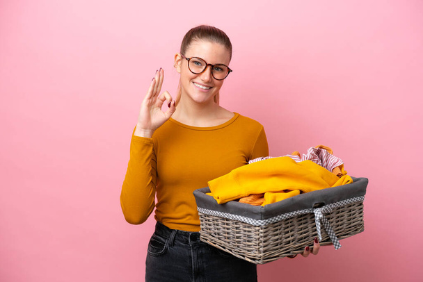 Young caucasian woman holding a clothes basket isolated on pink background showing ok sign with fingers - Foto, afbeelding