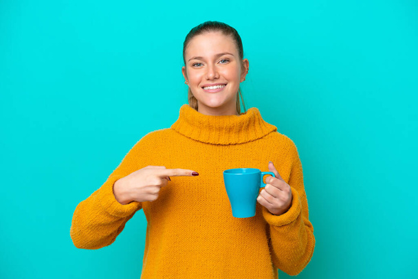 Young caucasian woman holding cup isolated on blue background and pointing it - Photo, Image