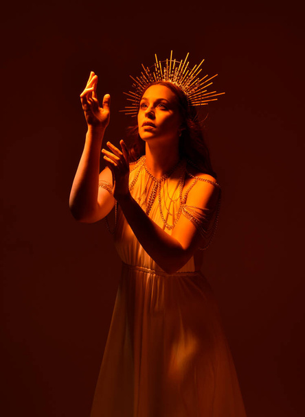 portret van mooie rode hoofd vrouw dragen lange vloeiende fantasie toga jurk met engelachtige gouden halo kroon op studio achtergrond - Foto, afbeelding