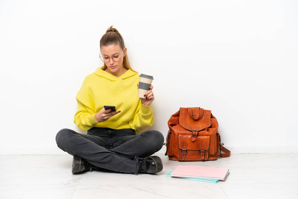 Young student caucasian woman sitting one the floor isolated on white background holding coffee to take away and a mobile - Photo, image