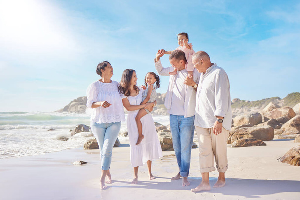 A smiling mixed race three generation family with little girls walking together on a beach. Adorable little kids bonding with mother, father, grandmother and grandfather outside. - 写真・画像