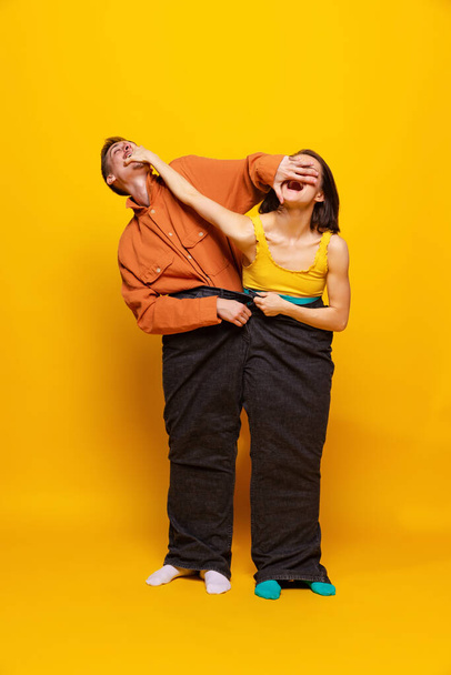 Kreatywny kolorowy portret mężczyzny i kobiety w jasnych ubraniach, stojących razem w jednych spodniach, pozujących na żółtym tle. Koncepcja mody retro, fotografii artystycznej, stylu, queer, piękna - Zdjęcie, obraz
