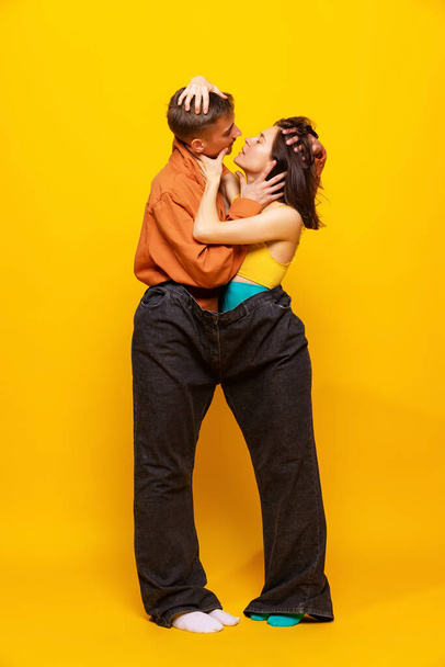 Kreatywny kolorowy portret mężczyzny i kobiety w jasnych ubraniach, stojących razem w jednych spodniach odizolowanych na żółtym tle. Miłości. Koncepcja mody retro, fotografii artystycznej, stylu, queer, piękna - Zdjęcie, obraz