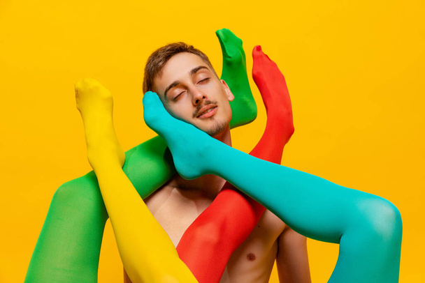 Creative colorful portrait of man with female legs in bright tights twining him isolated over yellow background. Extraordinary. Concept of retro fashion, art photography, style, queer, beauty - 写真・画像