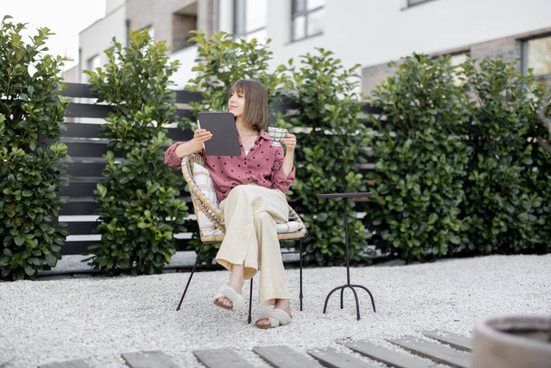 Giovane donna creativa lavora su tablet digitale mentre seduto rilassato con tazza di caffè nel cortile di casa sua. Concetto di lavoro a distanza o tempo libero con gadget all'aperto - Foto, immagini