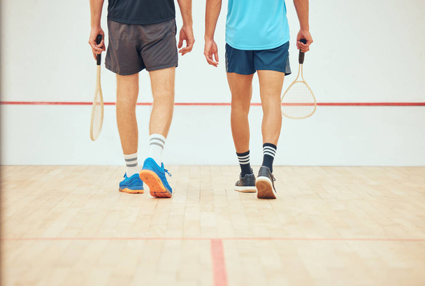 Rear view of two unknown squash players walking together on court and holding rackets before game. Fit active mixed race and caucasian athlete getting ready for training and practice in sports centre. - Photo, Image