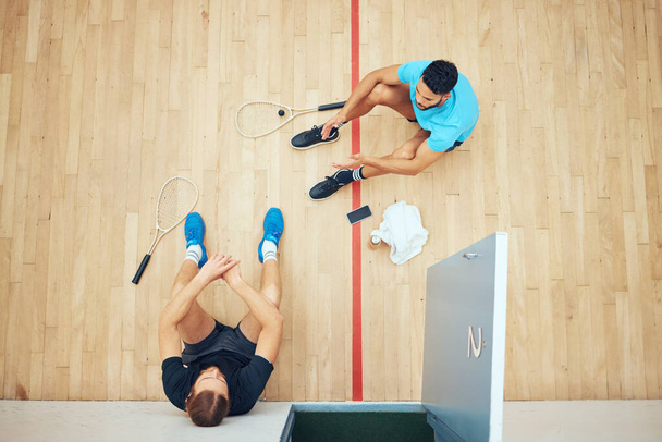 Above view of two unknown athletic squash players sitting together after playing court game. Fit active mixed race and caucasian athletes resting after training practice in sports centre. Sporty men. - Fotografie, Obrázek