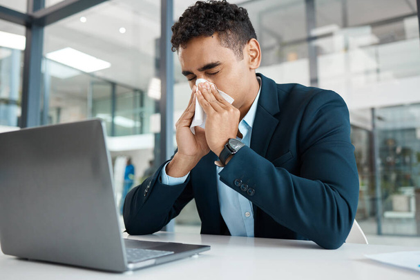 Sick mixed race businessman blowing his nose with a tissue while working on a laptop alone at work. One hispanic male businessperson suffering from allergies blowing his nose in an office. - Foto, Imagem