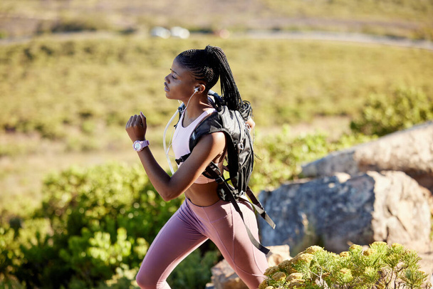 Ικανή και δραστήρια Αφρο-Αμερικανίδα γυναίκα που ακούει μουσική με ακουστικά, τρέχει μόνη της στα βουνά για προπόνηση. Εστιασμένη μαύρο αθλητή άσκηση και την κατάρτιση σε μονοπάτια στη φύση με ένα σακίδιο πλάτης - Φωτογραφία, εικόνα