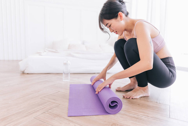 Young Asian woman wearing sportswear rolling yoga mat with water bottle, preparing for doing yoga. Working out at home or in yoga studio. Healthy habits, keep fit, weight loss concepts. - Photo, image
