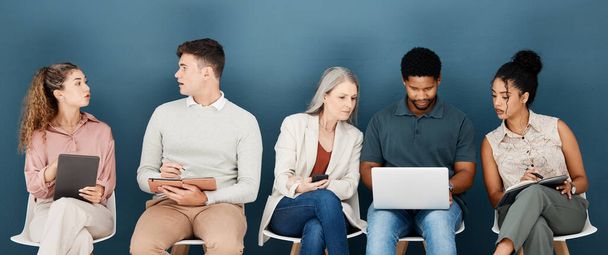 Group of diverse serious businesspeople sitting on chairs in a row in an office together. Five business professionals sitting and talking using technology in a waiting room together in an office. - Фото, зображення