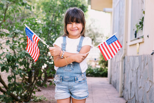 Malá tmavovlasá holčička s americkými vlajkami v zahradě svého domu. Koncepce oslav, Den nezávislosti, 4. července, vlastenectví, svátky a americká hrdost. - Fotografie, Obrázek