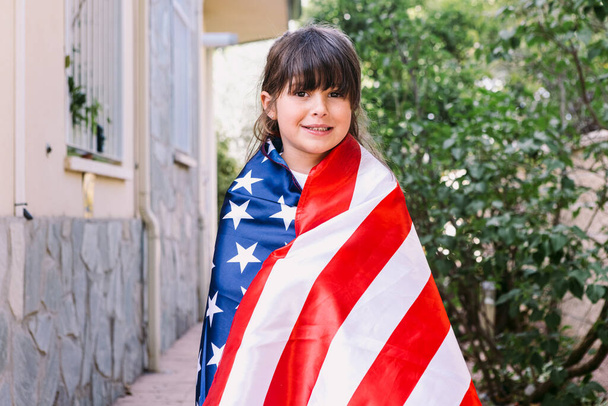 Fille aux cheveux noirs couverte d'un grand drapeau américain, dans le jardin de sa maison. Concept de célébration, fête de l'indépendance, États-Unis d'Amérique, 4 juillet, patriotisme et fierté américaine. - Photo, image