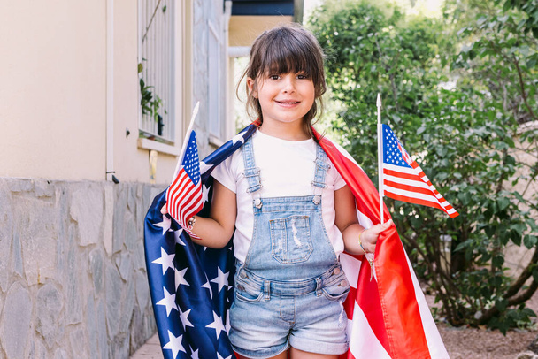 Piccola ragazza dai capelli neri coperta di grande bandiera degli Stati Uniti, con piccole bandiere americane nel suo giardino di casa. Concetto di celebrazione, giorno dell'indipendenza, 4 luglio, patriottismo, festa e orgoglio americano. - Foto, immagini