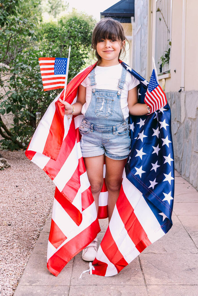 Pequena menina de cabelos pretos coberta com grande bandeira dos EUA, segurando pequenas bandeiras americanas em seu jardim em casa. Conceito de celebração, dia da independência, 4 de julho, patriotismo, feriado e orgulho americano. - Foto, Imagem