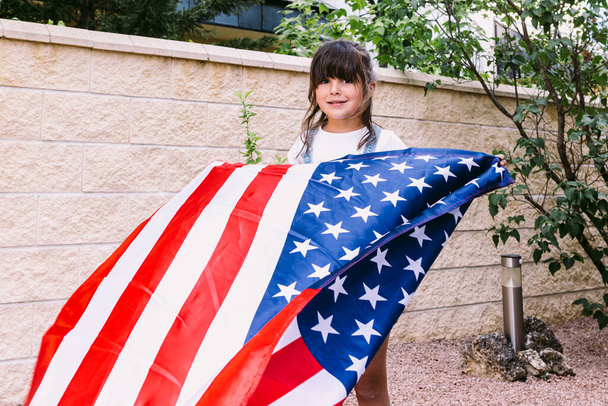 Schwarzhaariges Mädchen, das im Garten ihres Hauses eine US-Flagge hält und schwenkt. Konzept der Feier, Unabhängigkeitstag, Vereinigte Staaten von Amerika, 4. Juli, Patriotismus und amerikanischer Stolz. - Foto, Bild