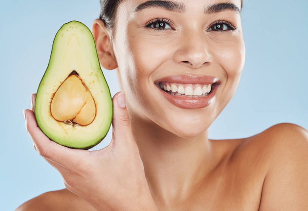 Beautiful young mixed race woman with an avocado isolated in studio against a blue background. Her skincare regime keeps her fresh. For glowing skin, eat healthy. Packed with vitamins and nutrients. - Photo, image