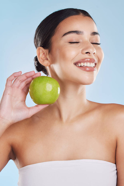 Beautiful young mixed race woman with an apple isolated in studio against a blue background. Her skincare regime keeps her fresh. For glowing skin, eat healthy. Packed with vitamins and nutrients. - 写真・画像