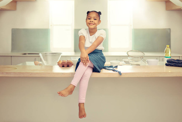 Portrait d'une jolie jeune fille métissée assise sur le comptoir de la cuisine avec un tablier et souriant. Petite fille hispanique souriante et assise seule tout en cuisinant à la maison. Elle aime cuisiner dans la cuisine. - Photo, image