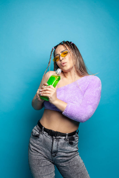 Cute young woman having fun with green plastic bottle in her hands against blue studio wall background - Foto, Imagem