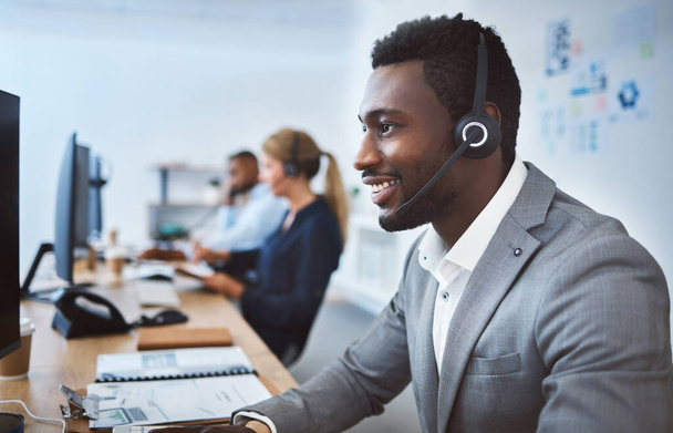 Happy young african american call centre telemarketing agent talking on a headset while working on computer in an office. Confident friendly male consultant operating helpdesk for customer service and - Φωτογραφία, εικόνα