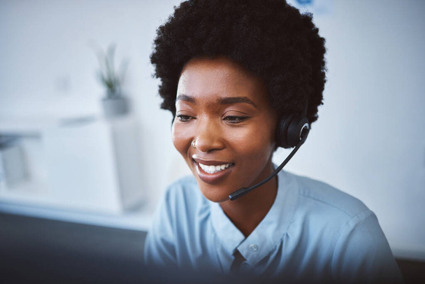 One happy young african american call centre telemarketing agent talking on a headset while working on computer in an office. Face of confident friendly female consultant operating helpdesk for - Foto, immagini