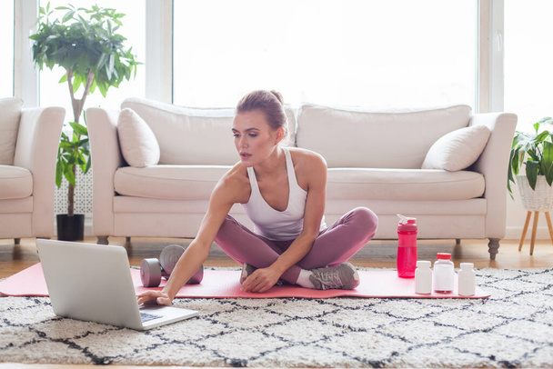 Mujer deportiva en ropa deportiva blanca y rosa sentada en el suelo con pesas y un batido de proteínas o una botella de agua y utilizando un ordenador portátil en casa en la sala de estar. Concepto de deporte y recreación. - Foto, imagen