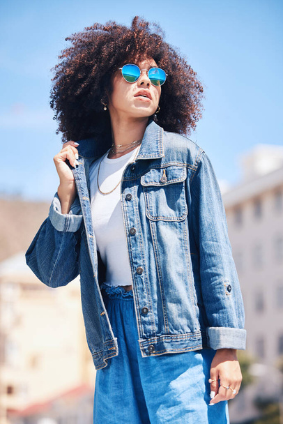 Cool and fashionable hispanic woman wearing sunglasses outside. Cheerful young mixed race woman with a curly afro wearing a trendy, stylish denim jacket while enjoying a summer day at the park outside - Zdjęcie, obraz