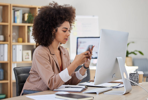 One young busy African American woman using a desktop and cellphone at a desk in her office job. Black woman with an afro checking social media at her job. Mixed race woman focused on texting. - 写真・画像
