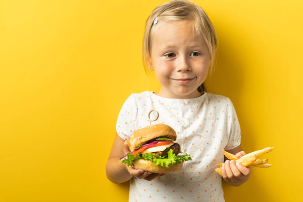 Cute little caucasian girl with blonde hair enjoying burger on a yellow background. Happy kid smiling and eating fast food burger - Fotografie, Obrázek