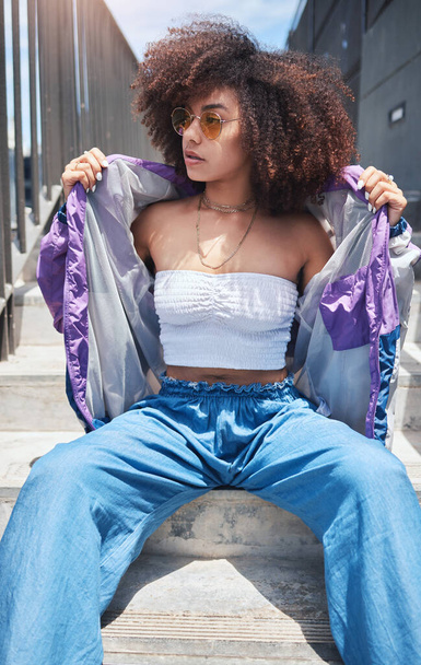 Young trendy beautiful mixed race woman with an afro sitting alone and posing alone outside. Hispanic woman with curly hair, sunglasses and hipster clothes. Fashionable African American woman in city. - Foto, immagini