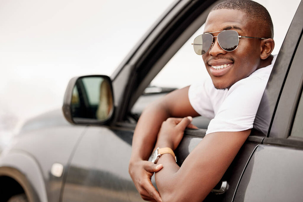 One African American man with his arm hanging out of the window, happy and looking back while taking a roadtrip. Black man enjoying the weekend and taking a trip in a vehicle while wearing sunglasses. - Fotó, kép