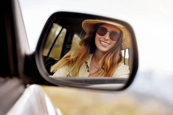 Smiling young caucasian woman looking at her reflection in the car mirror. Happy woman travelling, driving in her vehicle looking at herself in the mirror. Woman wearing sunglasses riding in a car. - Foto, Imagen