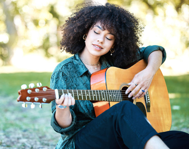 公園に座ってギターをひとりで演奏する中ぐりアフロを持つ若い混合レースの女性。若いです幸せなヒスパニック系の女性は、自然の中でリラックスしてギターを演奏しながら、. - 写真・画像