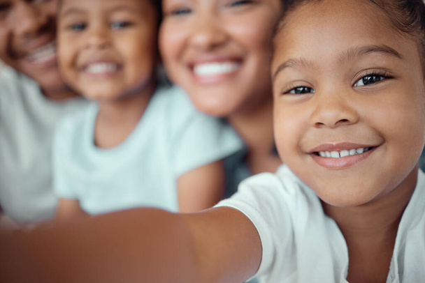 Close up of adorable little girl holding phone and taking selfie or recording funny video with her family. Happy mixed race family with two children and parents posing together for a family picture on - Foto, afbeelding