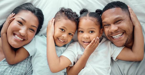 Portrait of happy mixed race family with two children relaxing and lying together on a bed at home, from above. Little brother and sister with their hands on each others faces while bonding with their - 写真・画像