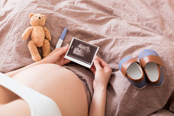 Kobieta w ciąży siedzi na łóżku i patrzy na zdjęcie USG swojego dziecka. Ciężarna samica pokazuje wyniki USG i nienarodzone dziecko.. - Zdjęcie, obraz