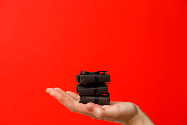 Mano femenina sosteniendo pequeñas cajas de regalo negras con cintas negras sobre fondo rojo intenso brillante - Foto, imagen