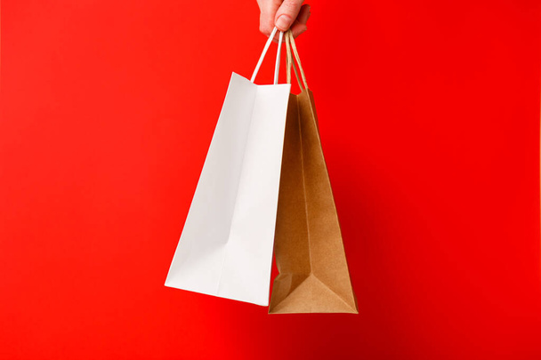 Weibliche Hand mit zwei Einkaufstaschen auf rotem Hintergrund. Weiße und braune Bastelpapiertüten in der Hand. Black-Friday-Verkauf, Discount, Shopping und Umweltkonzept - Foto, Bild