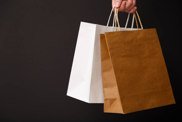 Female hand holding two shopping bags isolated on black background. White and brown craft blank paper bags in hand. Black friday sale, discount, shopping and ecology concept - Zdjęcie, obraz