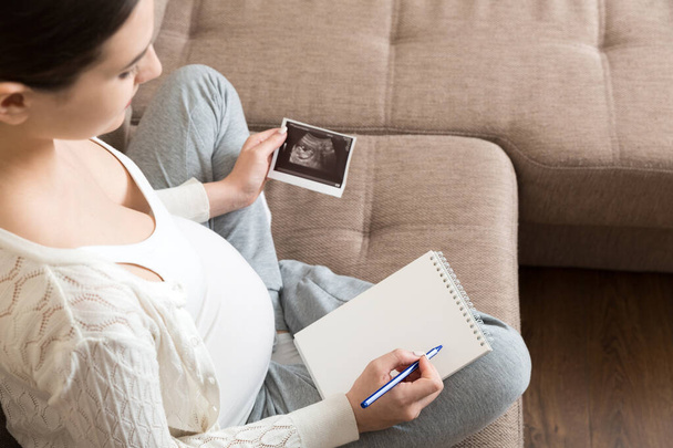 Top view opiekuńczy przyszłej matki piszącej pamiętnik z USG. kobieta w ciąży brzuch robi notatki uczucie podczas ciąży lub tworzenia albumu. - Zdjęcie, obraz