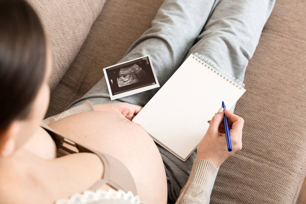 Top view caring future mother writing diary with ultrasound. pregnant woman tummy making notes feeling during pregnancy or creating scrapbook. - Photo, Image